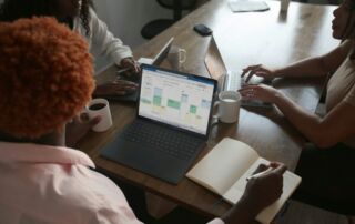 Three people in a meeting at a table discussing schedule on their Microsoft laptop